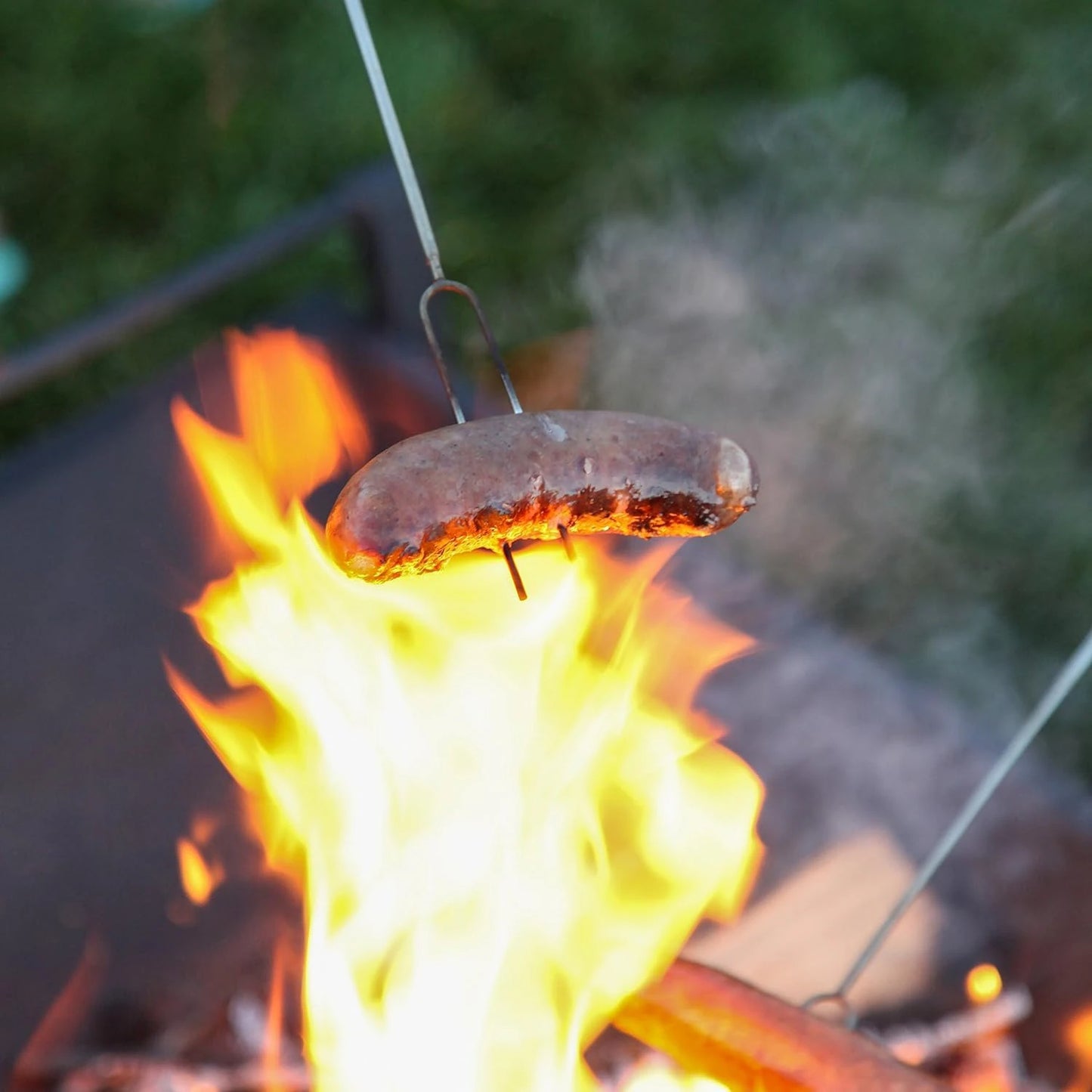 Telescoping s'mores roasting sticks with two prongs, roasting a sausage over a campfire. Perfect for camping and outdoor adventures.